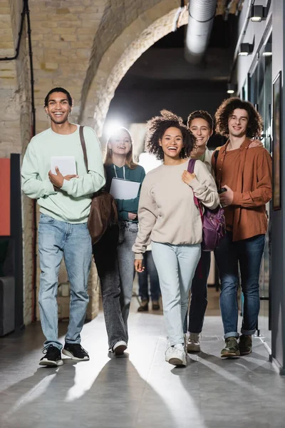 Pleine longueur de joyeux étudiants multiethniques marchant le long du couloir et regardant la caméra — Photo de stock