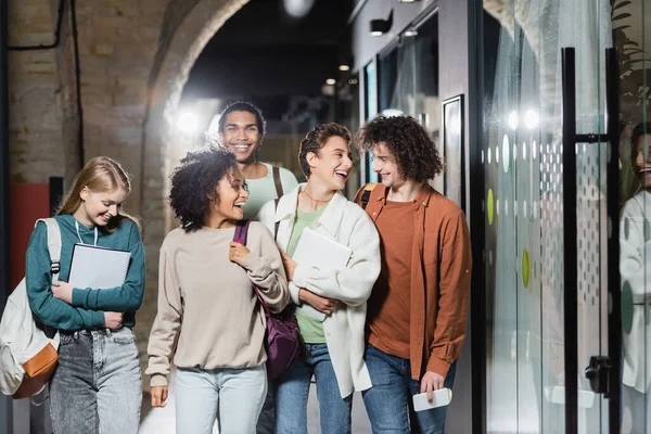 Jóvenes estudiantes multiétnicos con mochilas y gadgets sonriéndose en el pasillo - foto de stock