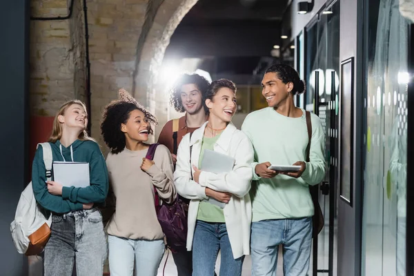 Cheerful multiethnic students smiling at each other while walking along corridor — Stock Photo