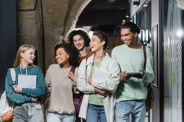 Jovens estudantes multiétnicos com gadgets e mochilas rindo no corredor — Fotografia de Stock