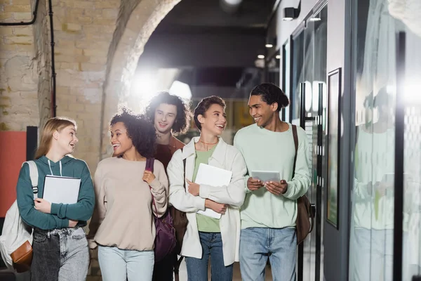 Estudiantes multiculturales felices con mochilas y gadgets caminando por el pasillo - foto de stock
