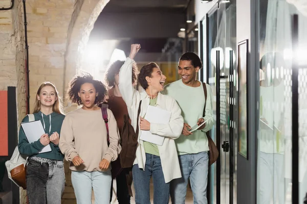 Mujer emocionada mostrando gesto de victoria cerca de amigos interracial en corredor universitario - foto de stock