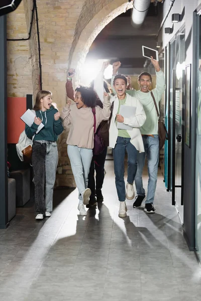 Longitud completa de estudiantes multiétnicos emocionados caminando por el pasillo y mostrando el gesto de ganar - foto de stock