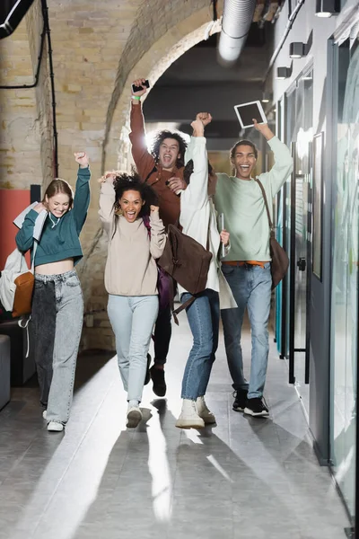 Estudiantes multiétnicos alegres con mochilas y gadgets que muestran el gesto de ganar en el corredor universitario - foto de stock