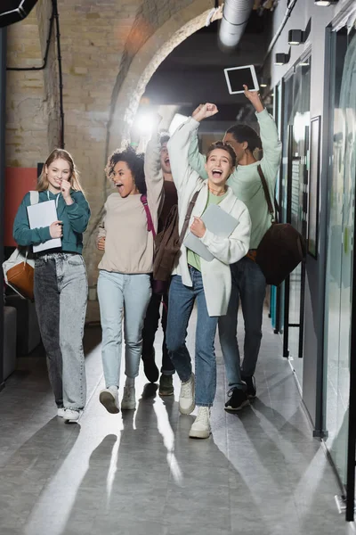 Full length of happy multicultural students showing success gesture while walking along corridor — Stock Photo