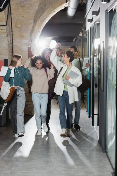 Amigos interracial emocionados caminando por el pasillo de la universidad y mostrando gesto de triunfo - foto de stock