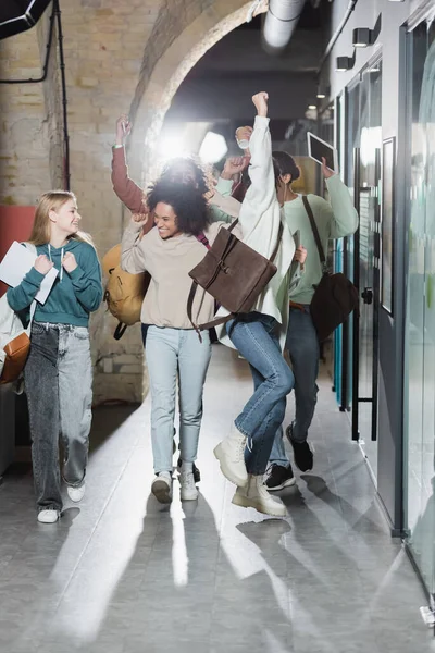 Fröhliche multiethnische Freunde, die den Flur der Universität entlanggehen und Jubelgeste zeigen — Stockfoto