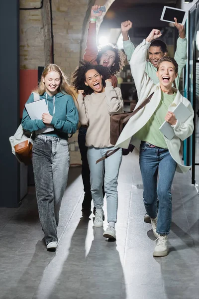 Happy multiethnic students screaming and showing win gesture while walking along hallway — Stock Photo