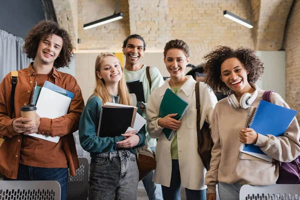 Heureux étudiants multiethniques avec copybooks et gadgets regardant la caméra dans l'auditorium universitaire — Photo de stock