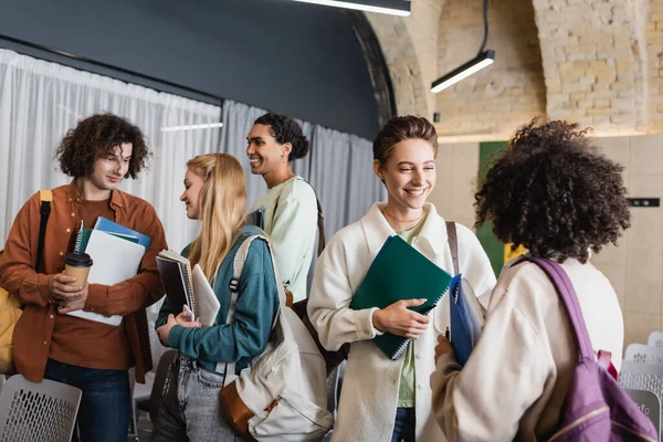 Estudantes multiétnicos alegres com copybooks e gadgets falando na universidade — Fotografia de Stock