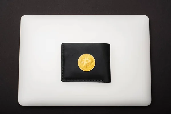 KYIV, UCRANIA - 26 DE ABRIL DE 2022: Vista superior de Bitcoin de oro en la cartera y el ordenador portátil aislado en negro - foto de stock