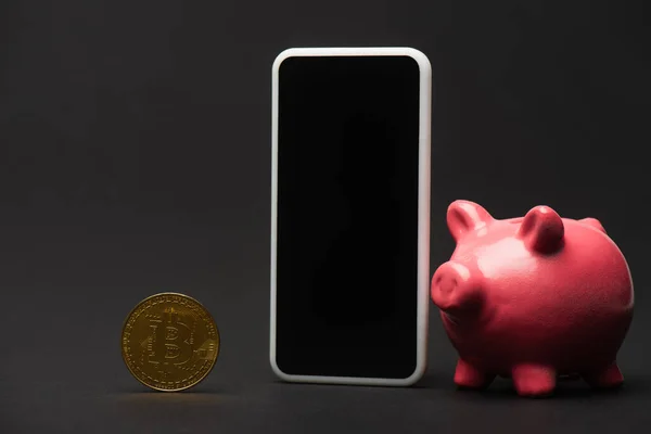 KIEW, UKRAINE - 26. April 2022: Nahaufnahme des Smartphones in der Nähe von Sparschwein und Bitcoin auf schwarzem Hintergrund — Stockfoto