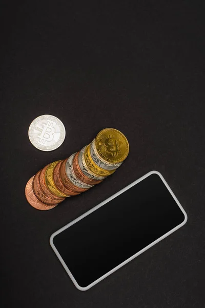 QUIIV, UCRÂNIA - 26 de abril de 2022: Vista superior de moedas criptografadas perto do smartphone com tela em branco no fundo preto — Fotografia de Stock