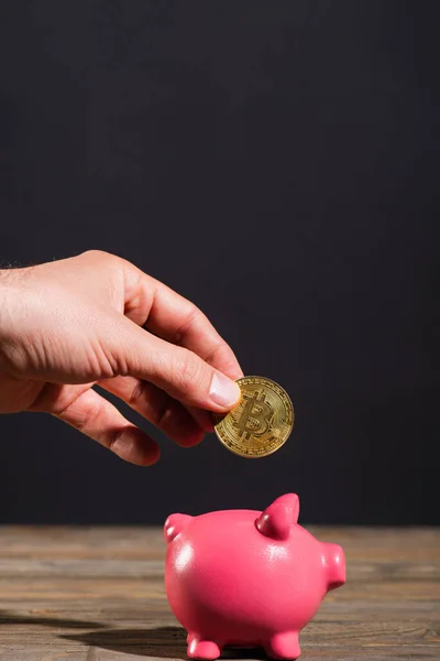 QUIIV, UCRÂNIA - ABRIL 26, 2022: Vista cortada do homem segurando bitcoin perto de banco porquinho na superfície de madeira isolada em preto — Fotografia de Stock