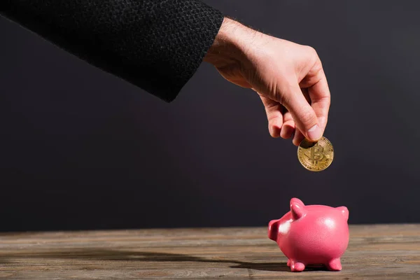 KYIV, UKRAINE - APRIL 26, 2022: Cropped view of man holding golden bitcoin near piggy bank on wooden surface isolated on black — Stock Photo