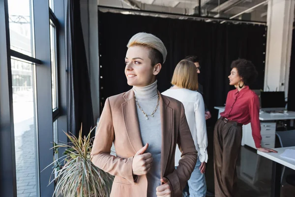 Feliz mujer de negocios mirando hacia otro lado cerca de las ventanas y borrosa equipo multicultural en agencia de publicidad - foto de stock