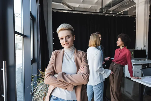 Glückliche Geschäftsfrau, die mit verschränkten Armen steht, während multiethnische Kollegen in Werbeagentur reden — Stockfoto