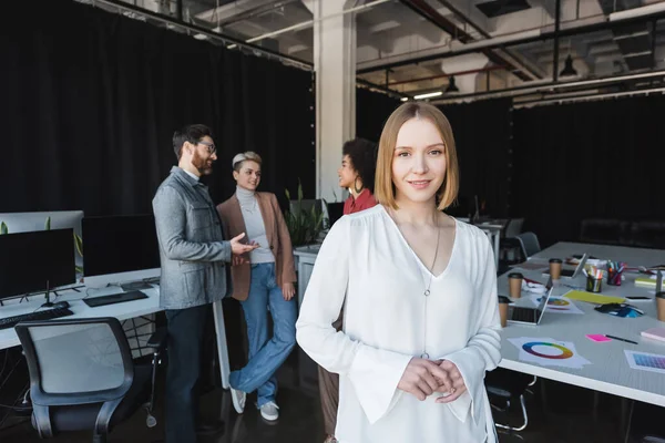 Lächelnde Geschäftsfrau blickt in die Kamera neben multikulturellen Kollegen, die auf verschwommenem Hintergrund reden — Stockfoto