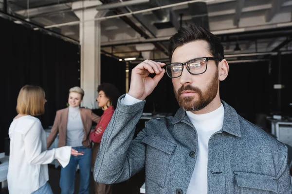 Hombre de negocios barbudo ajustando gafas y mirando hacia otro lado mientras el equipo multicultural habla sobre fondo borroso - foto de stock