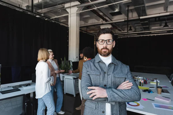 Advertising agent in eyeglasses standing with crossed arms near blurred multiethnic colleagues — Stock Photo