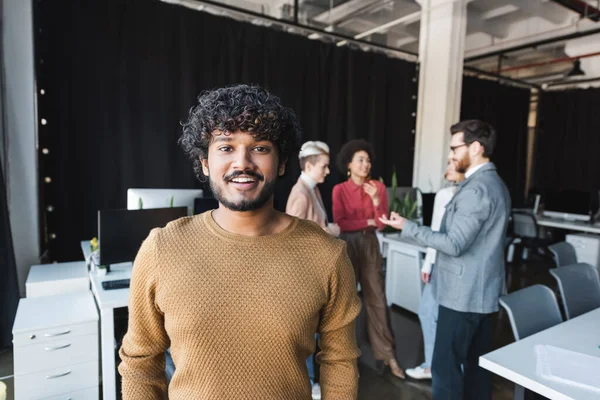 Bearded indian man smiling at camera near colleagues talking in ad agency — Stock Photo