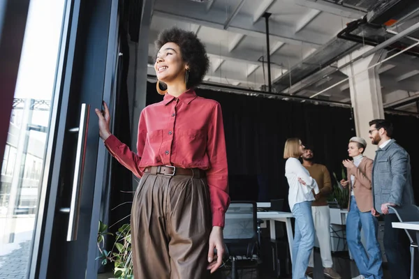 Vista de ángulo bajo del agente de publicidad afroamericano positivo mirando hacia otro lado cerca de colegas interracial en la oficina - foto de stock