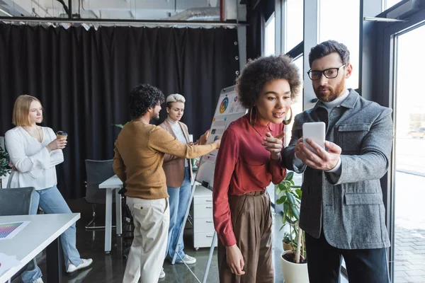 Businessman in eyeglasses showing smartphone to african american colleague in advertising agency — Stock Photo