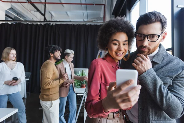 Donna afro-americana sorridente che mostra smartphone a collega premuroso in agenzia pubblicitaria — Foto stock