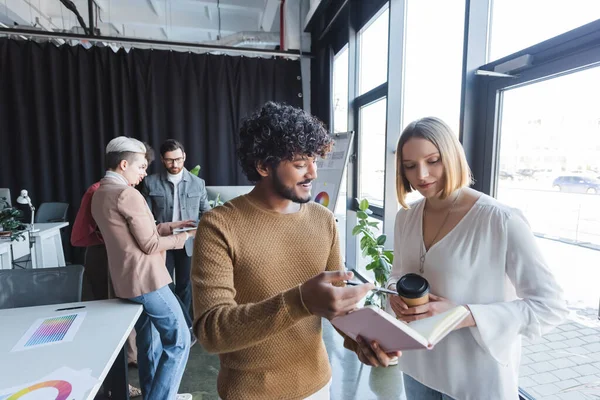 Hombre indio sonriente apuntando a un cuaderno cerca de un colega en la agencia de publicidad - foto de stock