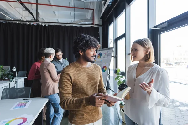 Woman with coffee to go talking to indian colleague with notebook in advertising agency — Stock Photo