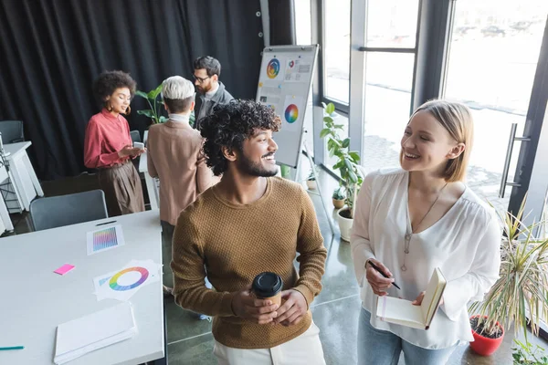 Cheerful interracial advertising agents with paper cup and notebook looking at each other in office — Stock Photo