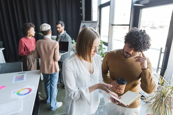 Woman pointing at notebook near thoughtful indian colleague in ad agency — Stock Photo