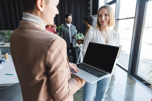 Mujer borrosa sosteniendo portátil con pantalla en blanco cerca de colega en la agencia de publicidad - foto de stock