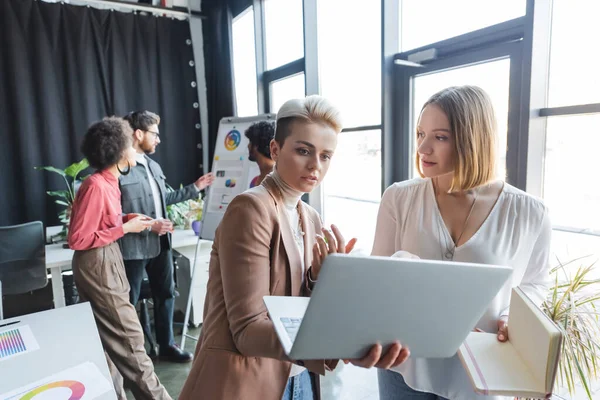Advertising managers talking near laptop and multiethnic colleagues near flip chart on background — Stock Photo