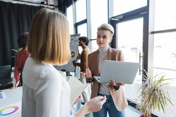 Woman holding laptop during conversation with blurred colleague in ad agency — Stock Photo