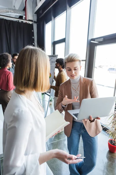 Lächelnder Werbemanager zeigt auf Laptop in der Nähe von Kollegen und interrassischen Mitarbeitern im Hintergrund — Stockfoto