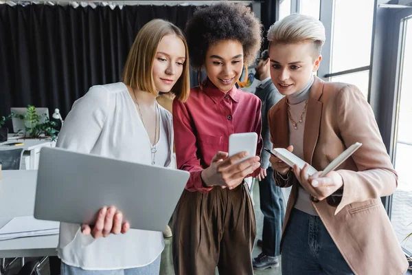 Gerentes de publicidad multiétnicos positivos con gadgets y cuaderno en la oficina - foto de stock