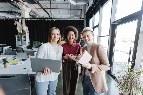 Alegres mujeres multiétnicas con portátil y gadgets mirando a la cámara en la agencia de publicidad - foto de stock