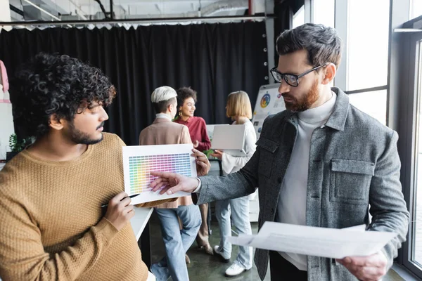 Человек в очках, указывающий на образцы цветов рядом с индийским дизайнером в рекламном агентстве — стоковое фото