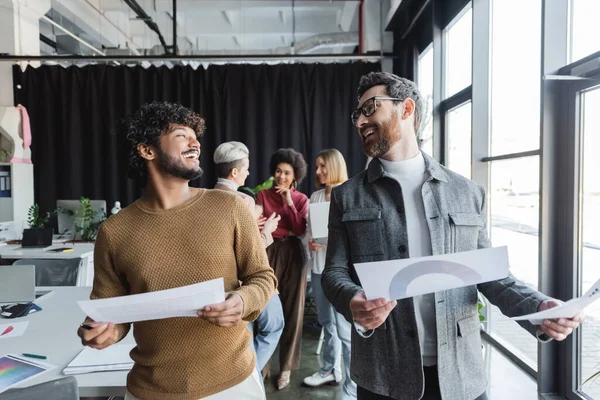 Hombres interracial riendo con papeles mirándose unos a otros en la agencia de publicidad - foto de stock