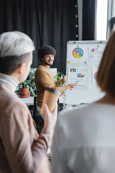 Happy indian advertising designer talking to blurred colleagues near flip chart with graphs — Stock Photo