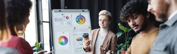 Donna con caffè per andare a parlare con colleghi interrazziali offuscati vicino lavagna a fogli mobili in agenzia pubblicitaria, banner — Foto stock