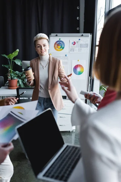 Smiling woman with coffee to go talking to blurred colleague in advertising agency — Stock Photo