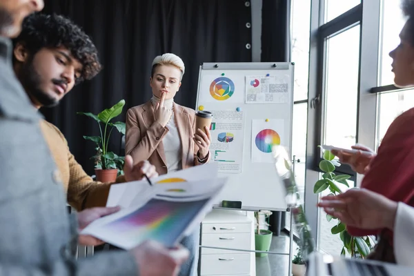 Thoughtful woman with coffee to go near blurred interracial team in advertising agency — Stock Photo