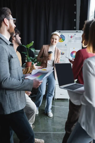 Cheerful advertising manager standing with coffee to go near blurred interracial colleagues — Stock Photo