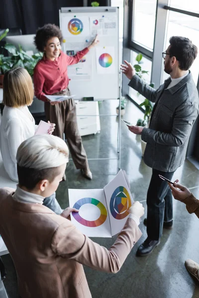 Smiling african american manager pointing at flip chart with project near colleagues in advertising agency — Stock Photo