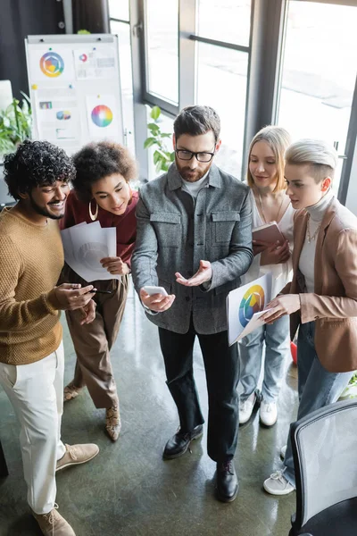 Man in eyeglasses pointing at smartphone near interracial colleagues in advertising agency — Stock Photo