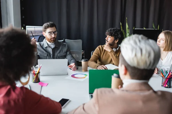 Uomo in occhiali che tiene la penna mentre parla con colleghi multietnici durante l'incontro in agenzia pubblicitaria — Foto stock