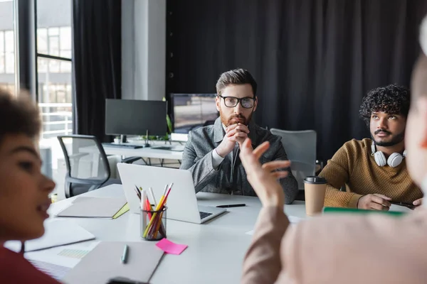 Bearded man in eyeglasses sitting with clenched hands during meeting with multiethnic advertising agents — Stock Photo