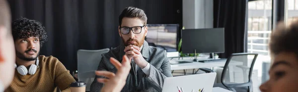 Thoughtful man in eyeglasses near indian colleagues and blurred people in advertising agency, banner — Stock Photo